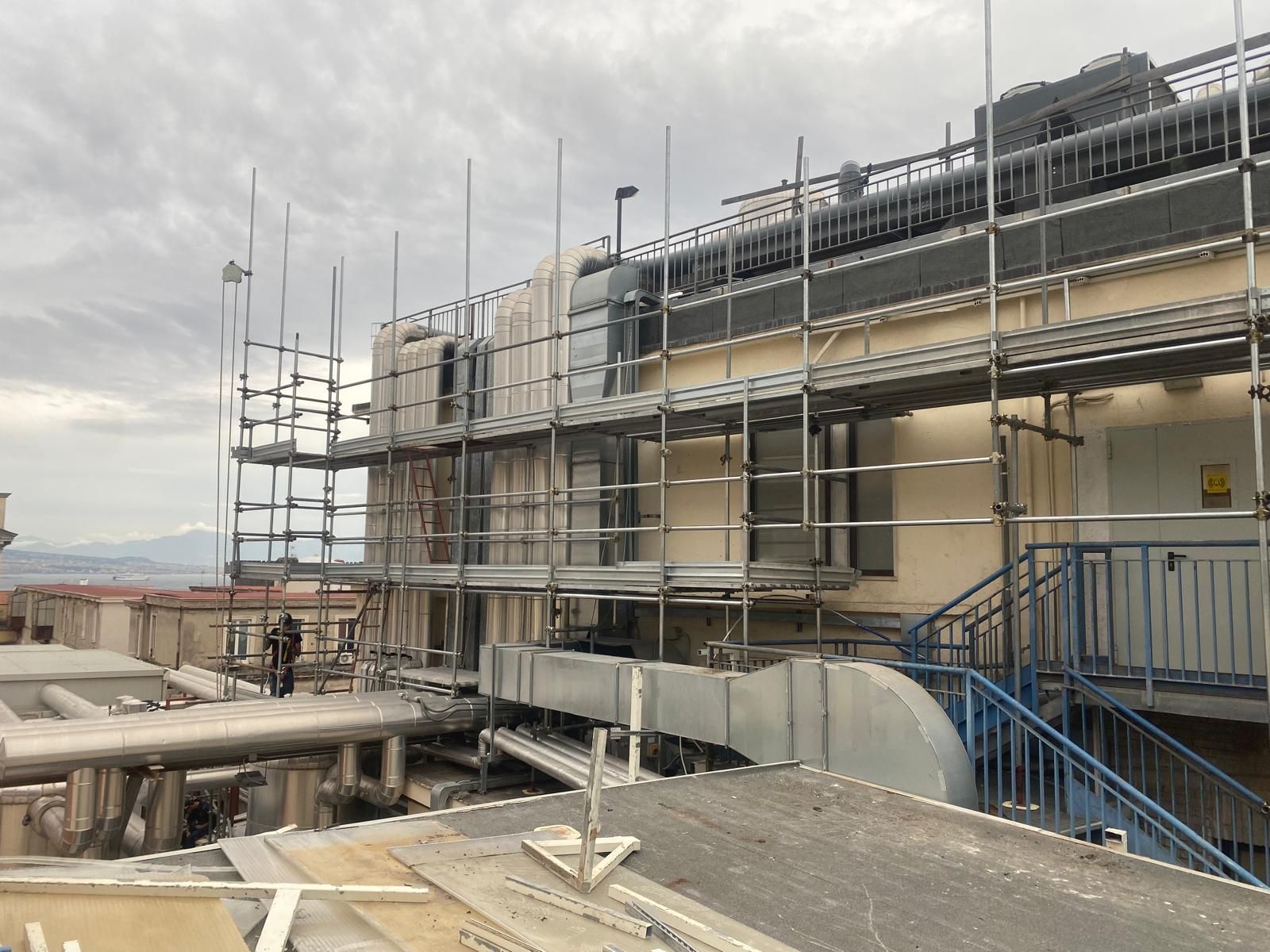 Un'impalcatura circonda l'esterno di un edificio. Delle scale blu conducono in alto. Il cielo è coperto.