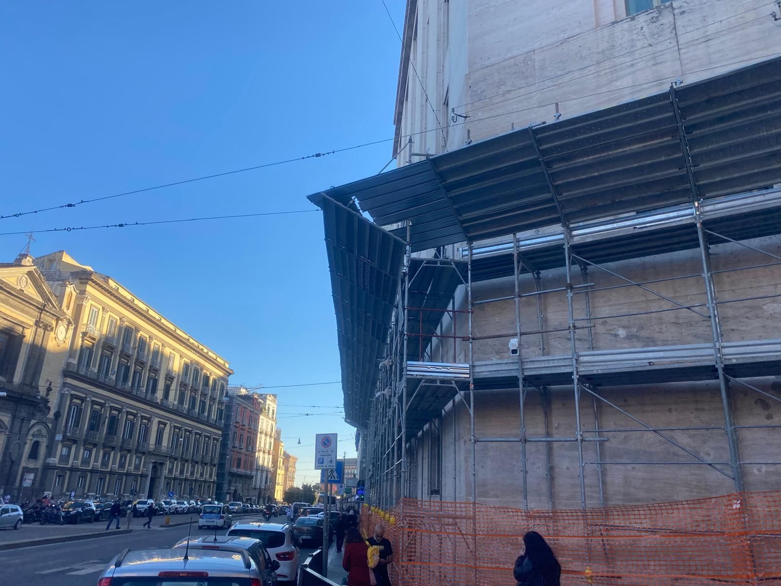 Scena di strada con impalcature all'angolo di un edificio; sono visibili automobili, persone e altri edifici.