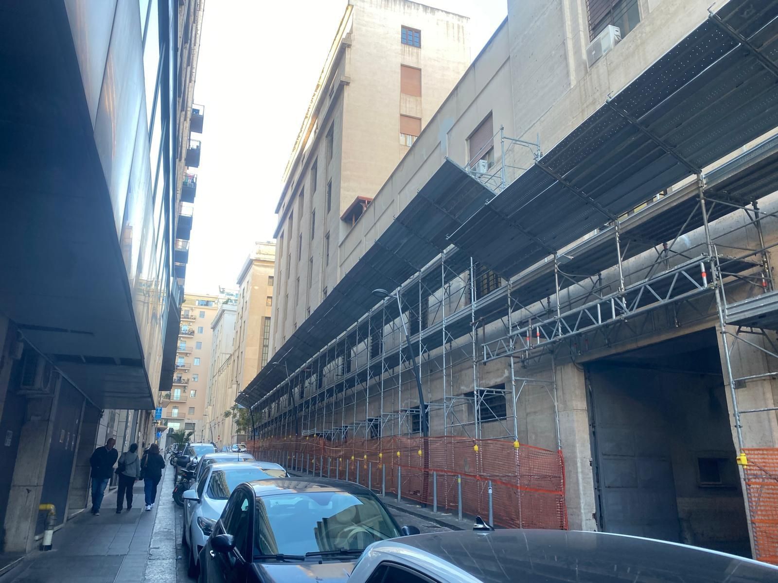Stretta strada cittadina con auto parcheggiate, pedoni e impalcature lungo un alto edificio.