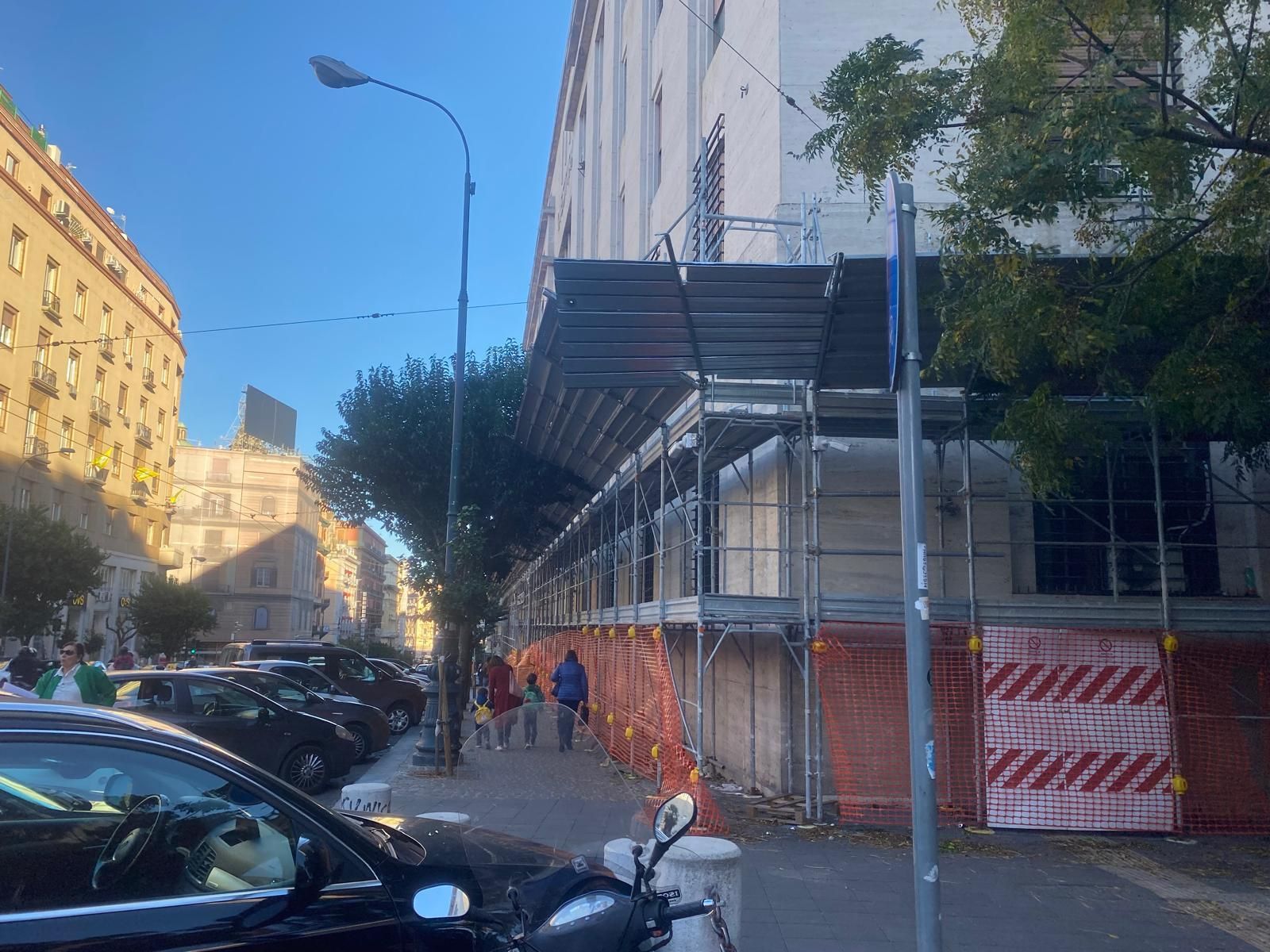 Un'impalcatura circonda un edificio in una strada cittadina. Sono visibili auto e pedoni.