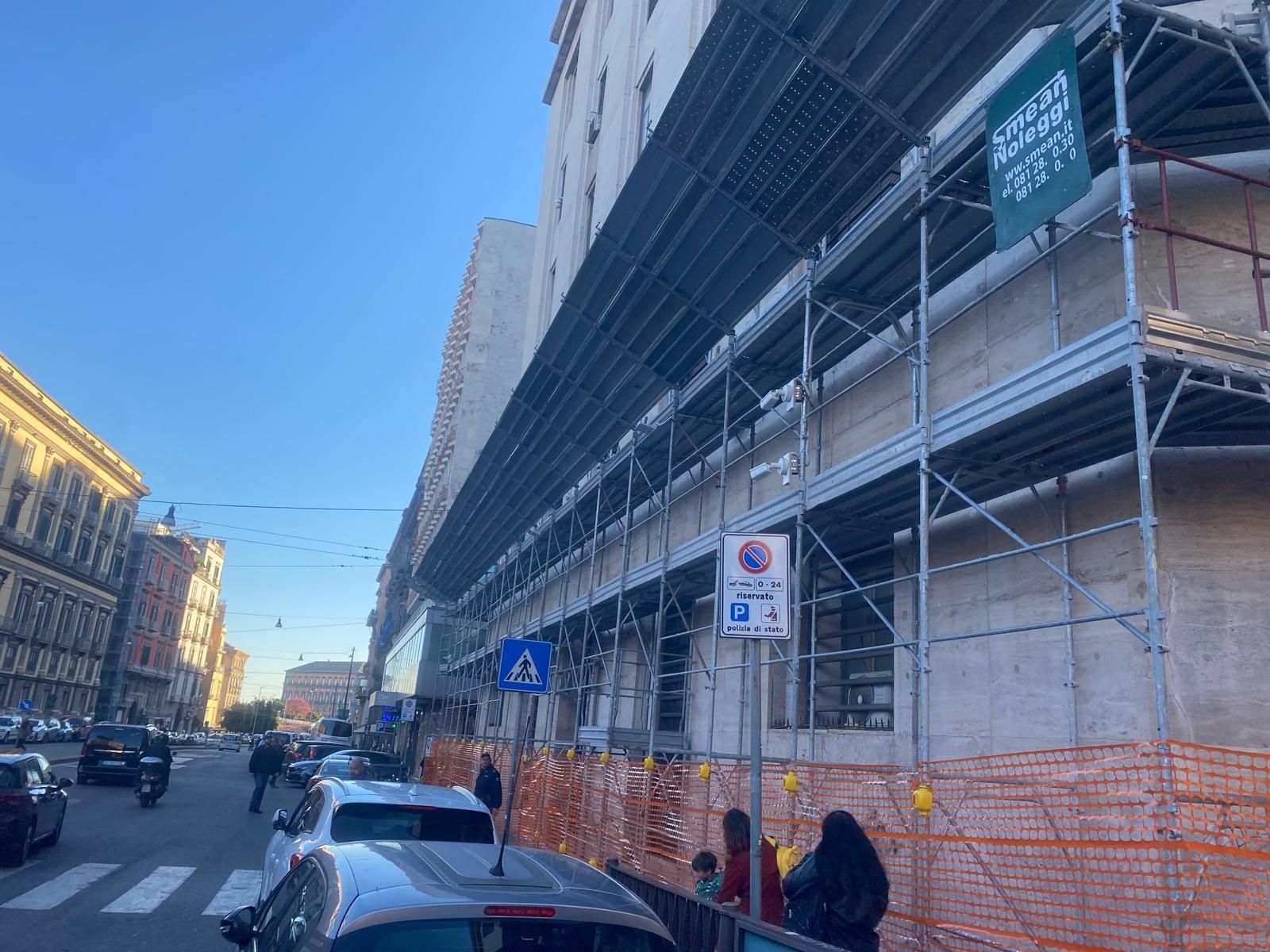 Impalcature sulla facciata di un edificio. Scena di strada con auto parcheggiate, pedoni e cielo azzurro.