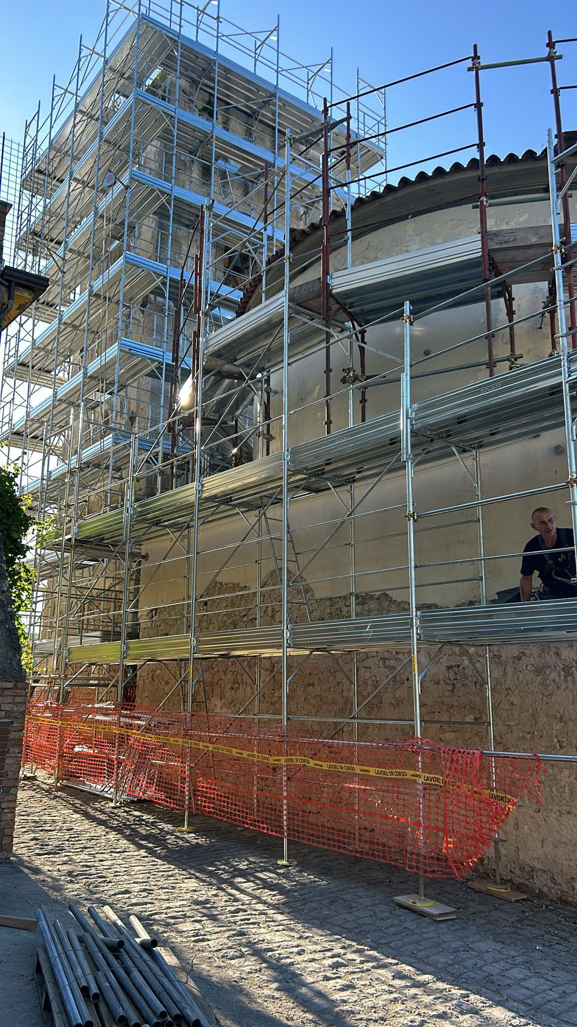 Un'impalcatura è stata eretta contro un edificio, con una rete di sicurezza arancione. Una persona è seduta sul tetto.