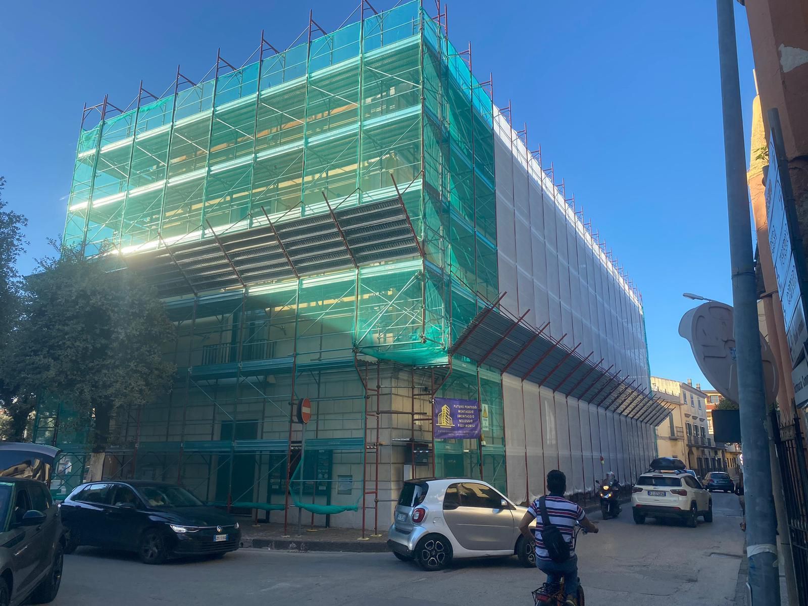 Edificio in costruzione, avvolto da impalcature e reti di sicurezza verdi su una strada cittadina; auto parcheggiate nelle vicinanze.