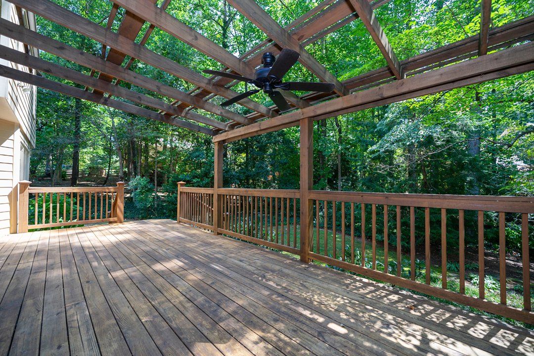 An empty wooden deck with a ceiling fan and trees in the background.