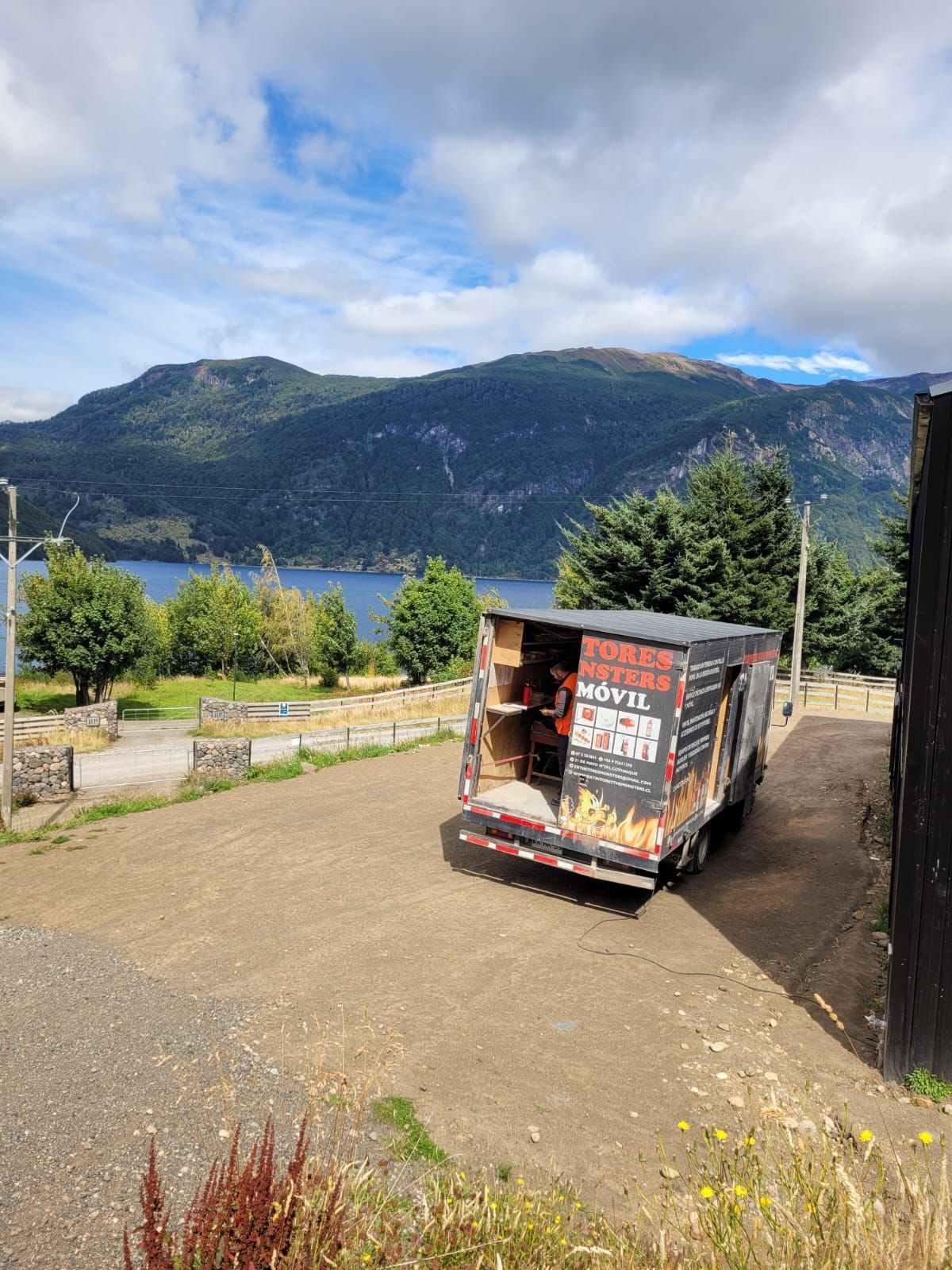 Remolque estacionado en una ladera con un lago y montañas al fondo. Cielo nublado.