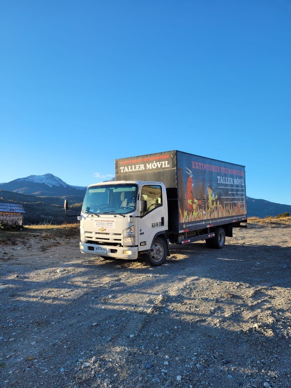 Camión blanco estacionado sobre grava, con montañas al fondo y un cielo azul. Un lateral tiene un anuncio.