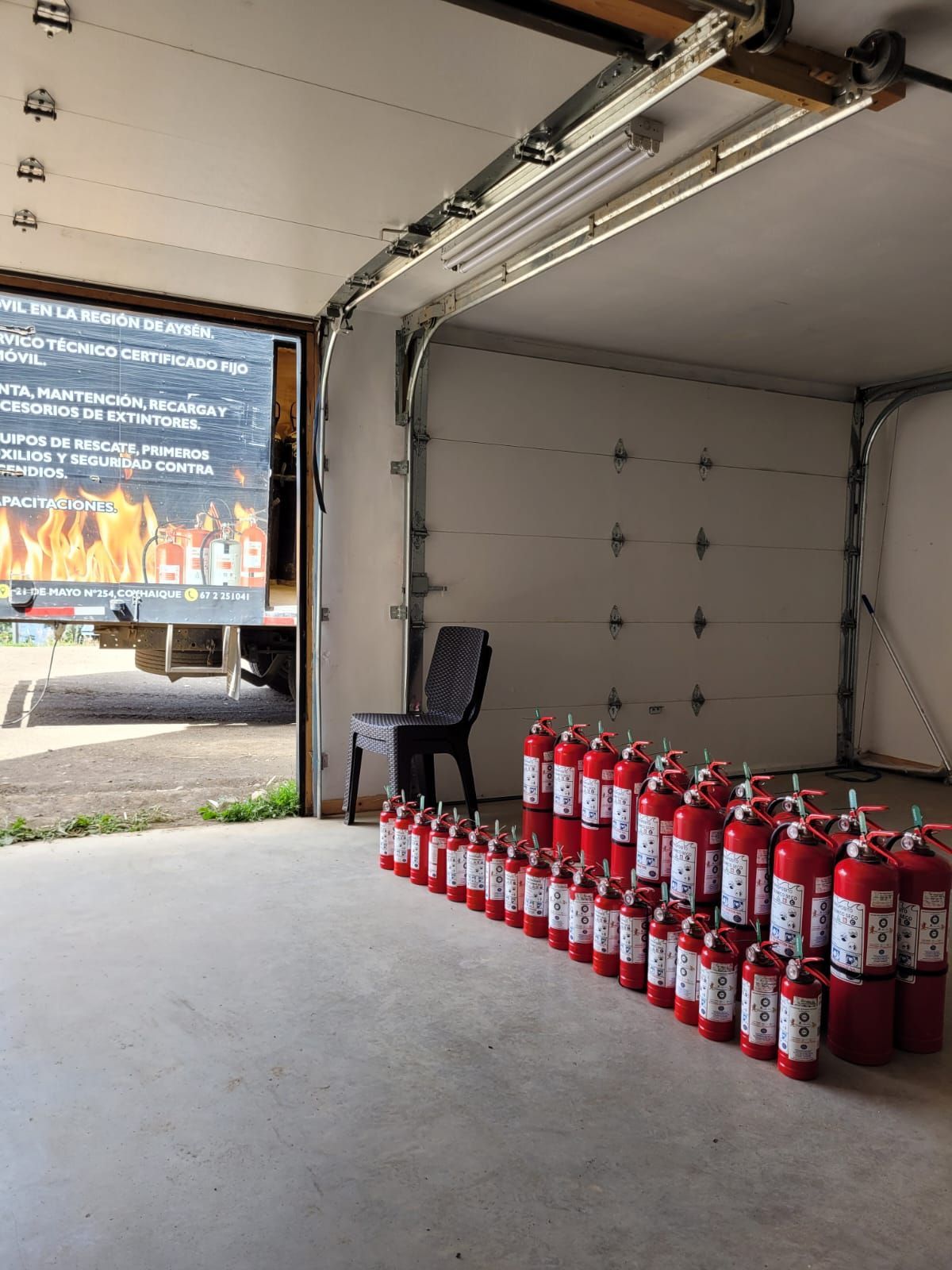 Extintores rojos dispuestos en un garaje; hay una silla cerca y afuera se ven un camión y un cartel.
