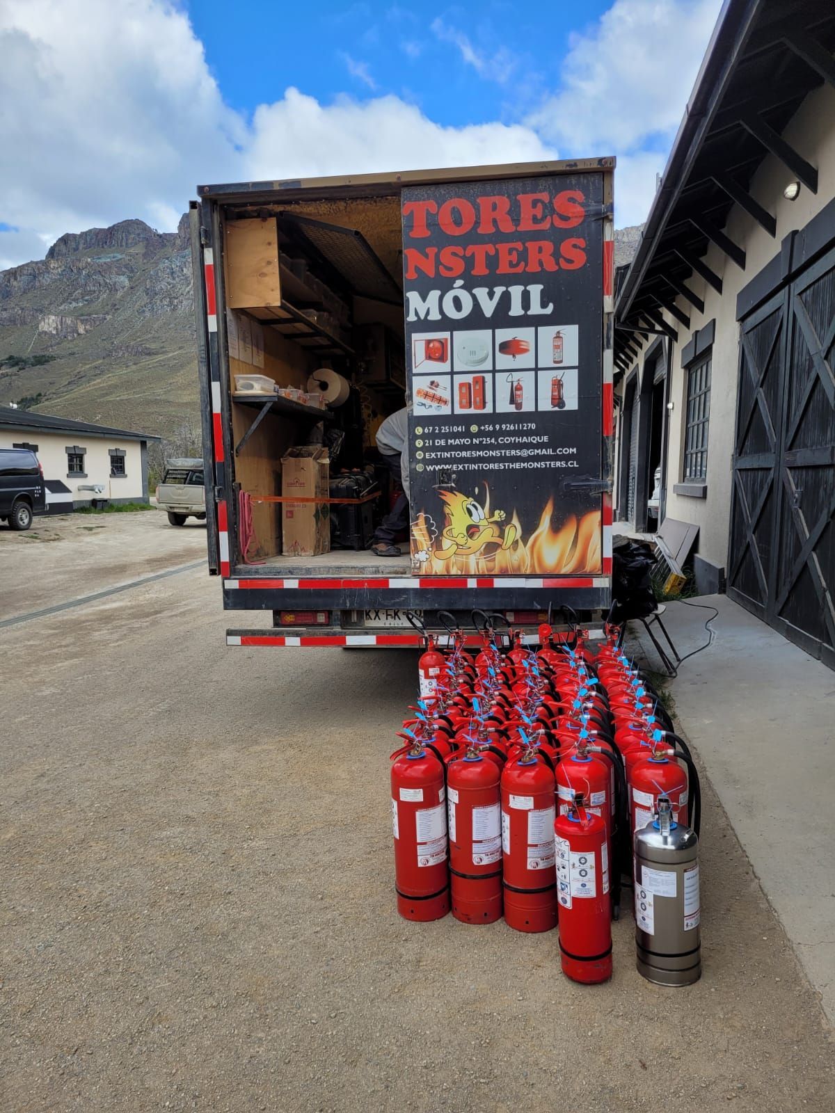 Extintores delante de un camión, probablemente para la venta o el servicio técnico. Edificio y montañas al fondo.