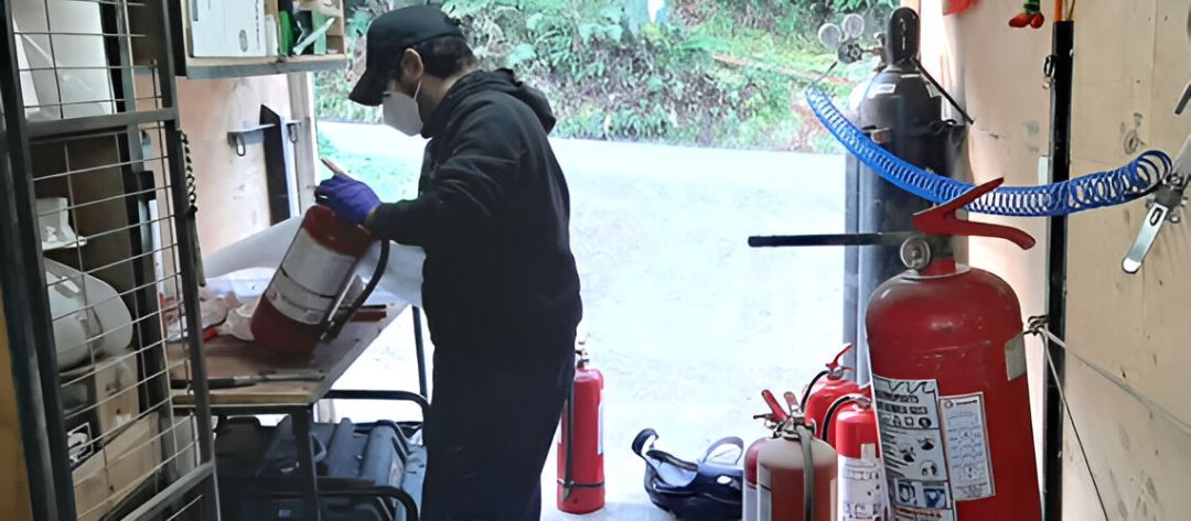 Persona con mascarilla y guantes realizando el mantenimiento de un extintor rojo en un taller.