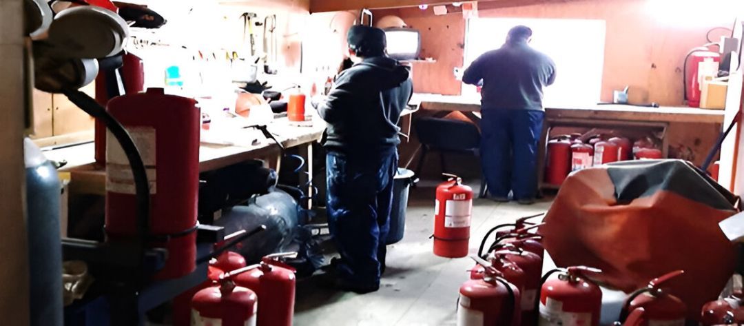 Dos hombres trabajando en un taller desordenado, lleno de extintores rojos. Uno junto a la ventana, el otro en un banco de trabajo.