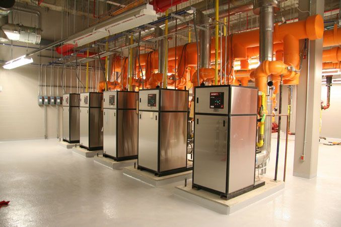 Row of industrial silver mechanical units with orange pipes, in a room with a white floor and grey walls.