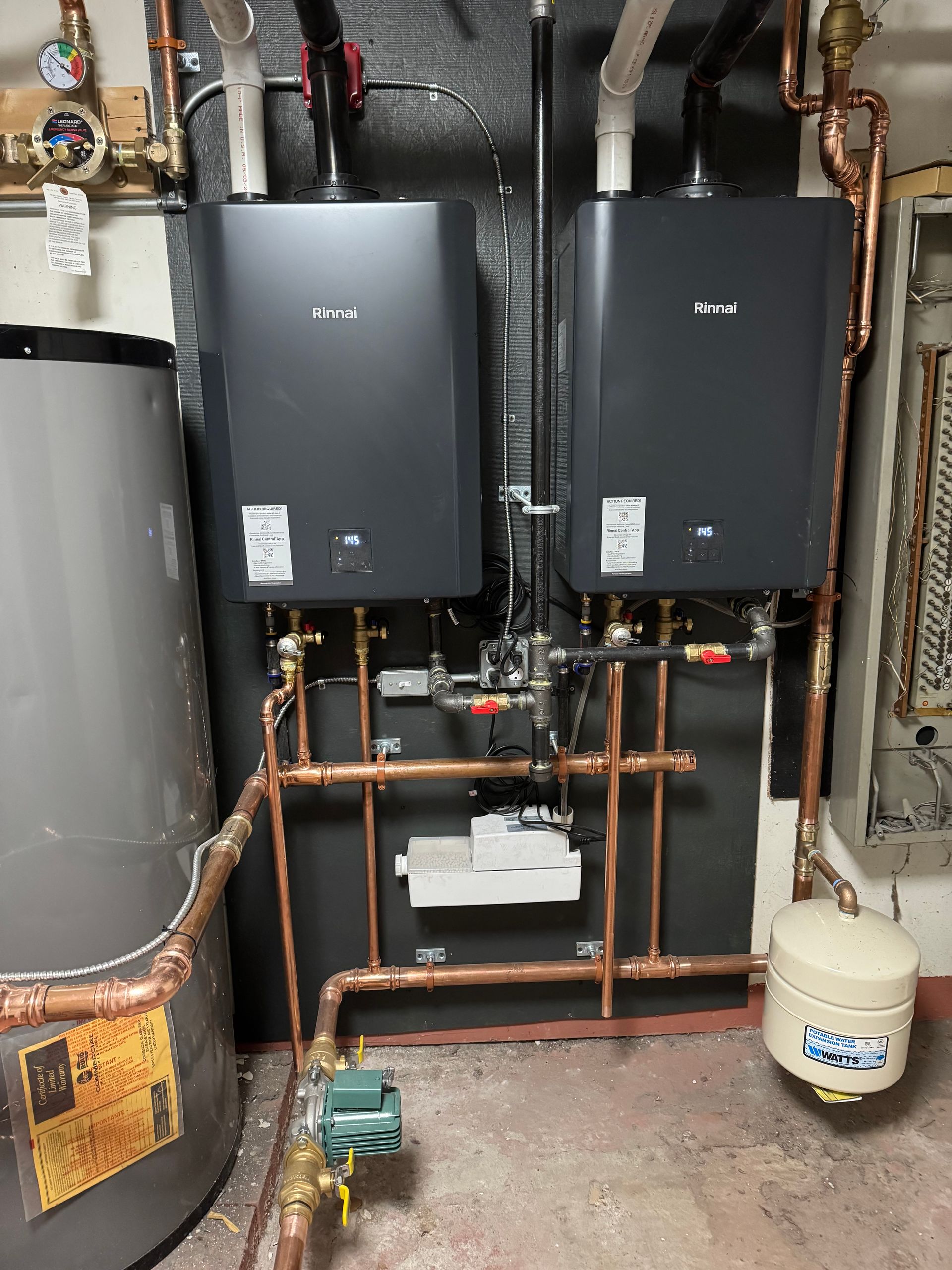 Two black wall-mounted boilers with copper pipes and a water tank, in a utility room.