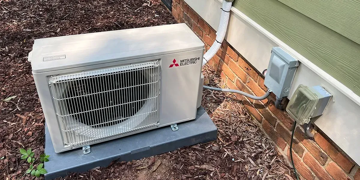 A Mitsubishi heat pump unit sits on a concrete base beside a brick wall. Power connections are visible.