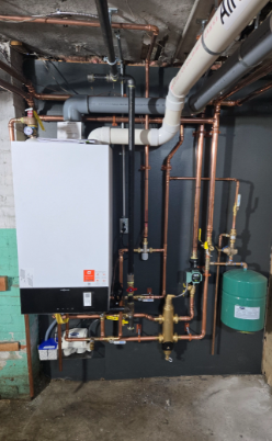 A boiler unit with copper pipes and a green expansion tank against a gray painted wall.