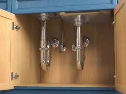 Under-sink plumbing with shiny chrome pipes and drain connections in a light wood cabinet with blue trim.