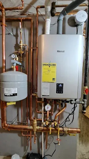 Copper-piped home heating system with a silver water heater, expansion tank, and gauges against a concrete wall.