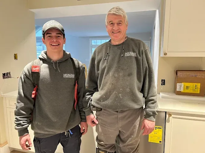 Two men in gray sweatshirts stand inside a kitchen. One young, one older. Cabinets and unfinished walls visible.
