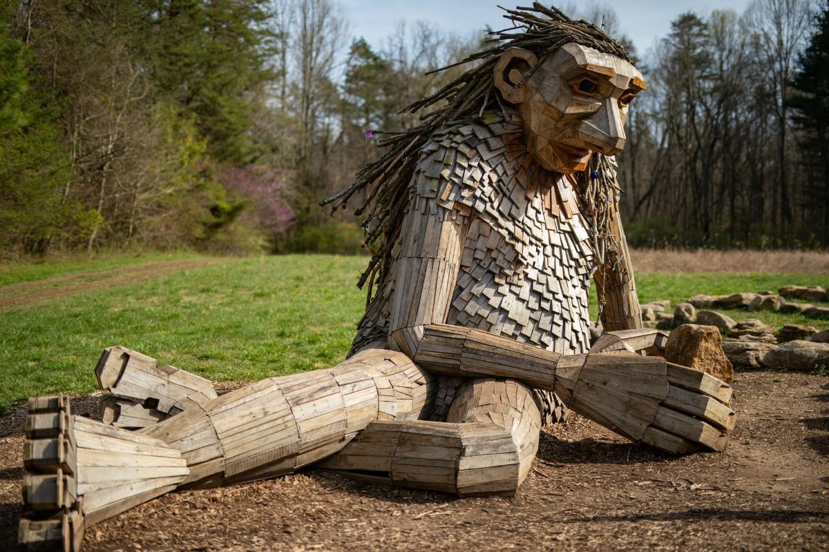 A large wooden statue of a troll is sitting on the ground in a field.