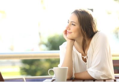 Mulher com a mão no queixo, sorrindo, sentada a uma mesa ao ar livre com uma xícara de café.