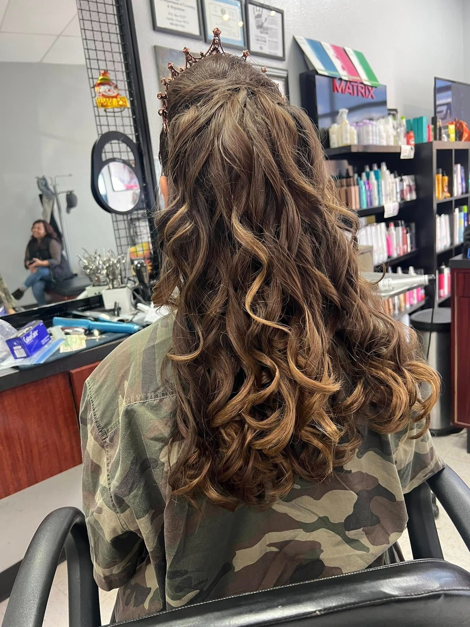 Person with styled, curly brown hair in a salon chair, wearing a camo jacket.