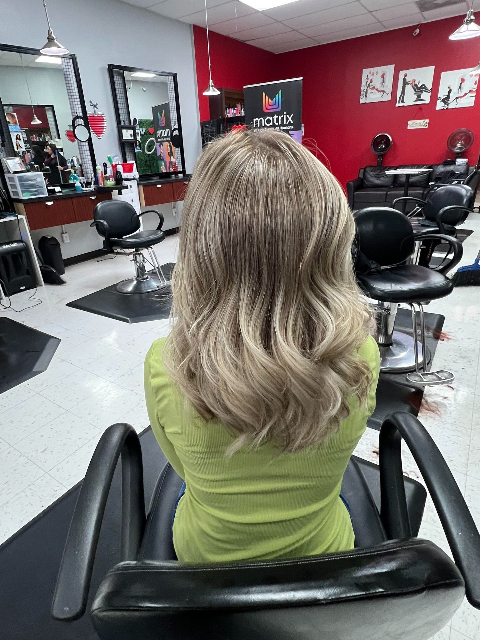 Woman in green shirt sits in a salon chair with wavy blonde hair, interior shot.