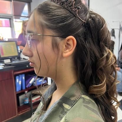 Young person with curly ponytail, glasses, and crown. Indoors, wearing a camouflage jacket.
