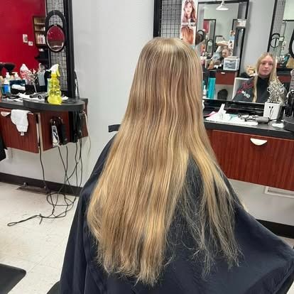 Person with long blonde hair in a salon chair, facing away. Hair is draped over a black cape. Salon setting.