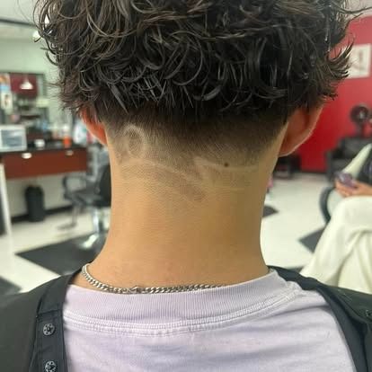 Person with short, curly hair and a fade haircut. Decorative design shaved into the nape of the neck, inside a barbershop.