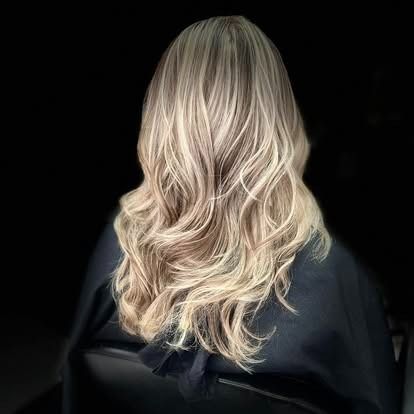 Woman with long, wavy blonde and brown highlighted hair, sitting in a black chair, wearing a black salon cape.