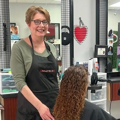 Woman in glasses and apron, cutting a person's curly hair in a salon.