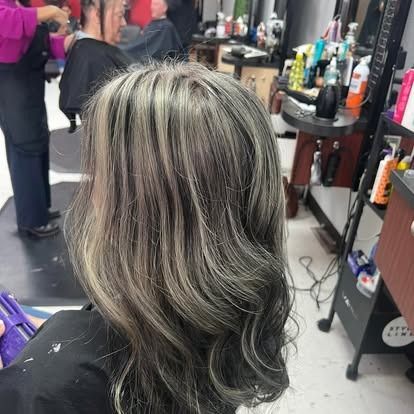Person with gray and blonde highlights in hair, at a salon. Other people and equipment are visible in the background.