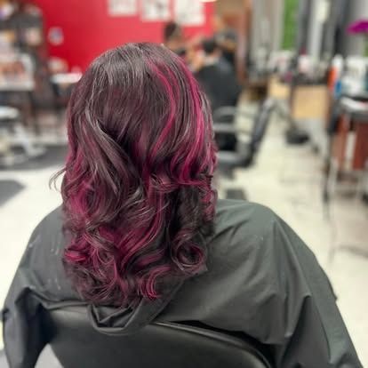 Woman in salon chair with dark hair, streaks of bright pink, and layered waves.