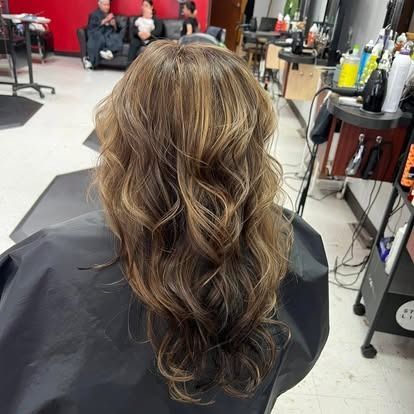Woman with wavy brown hair and highlights at a salon, wearing a black cape.