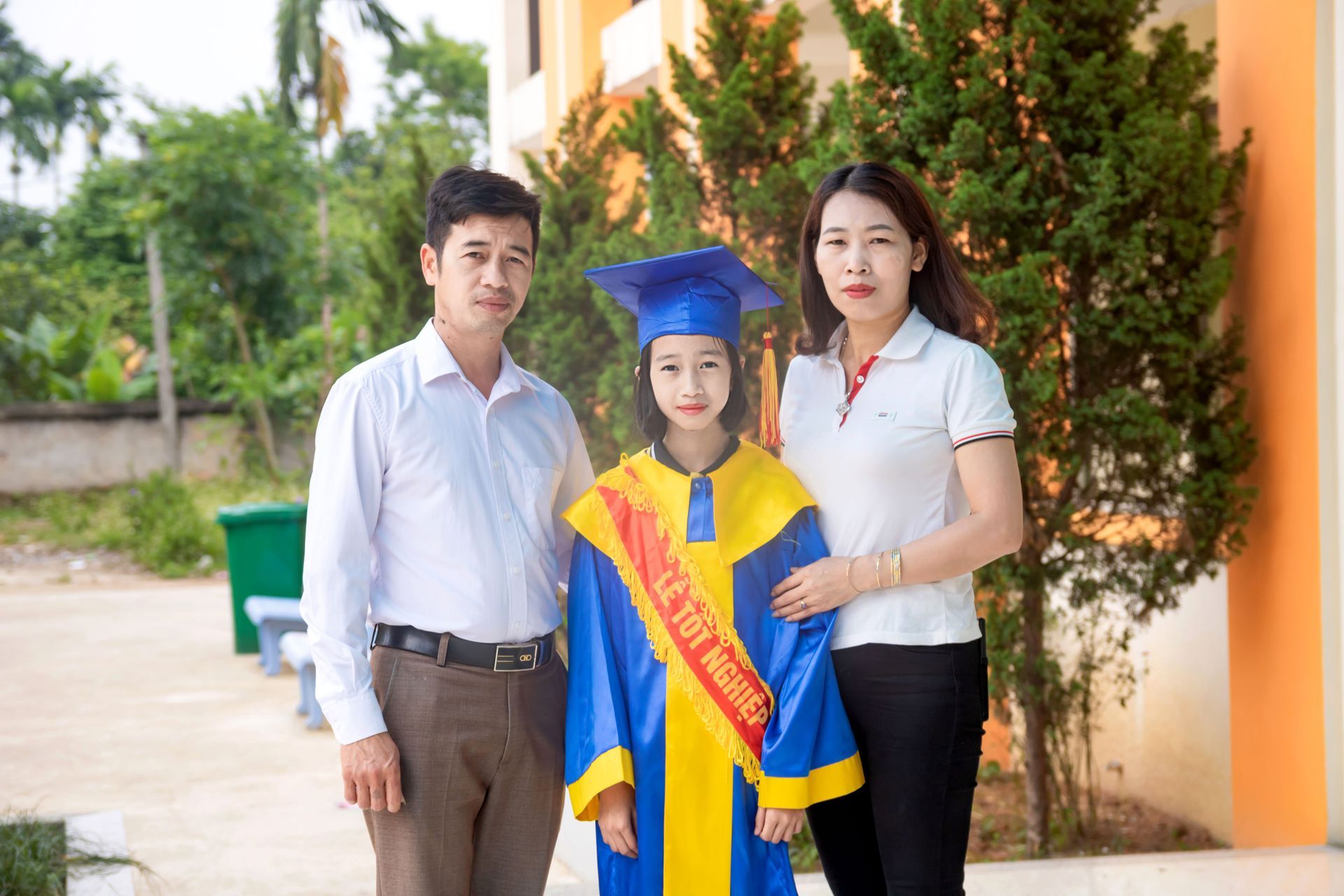Graduate in blue cap and gown stands with two adults outdoors by a building.
