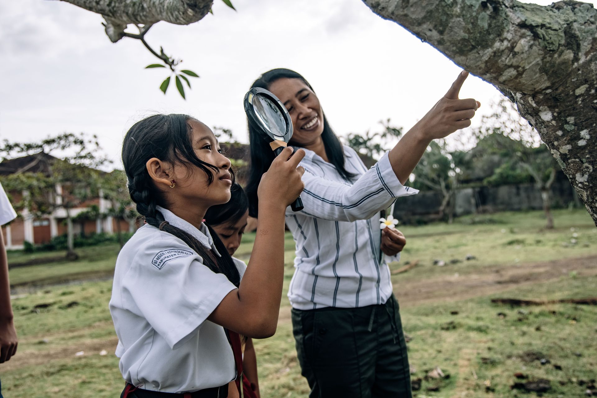 Two students in white uniforms outdoors, one using a magnifying glass and the other pointing upward.