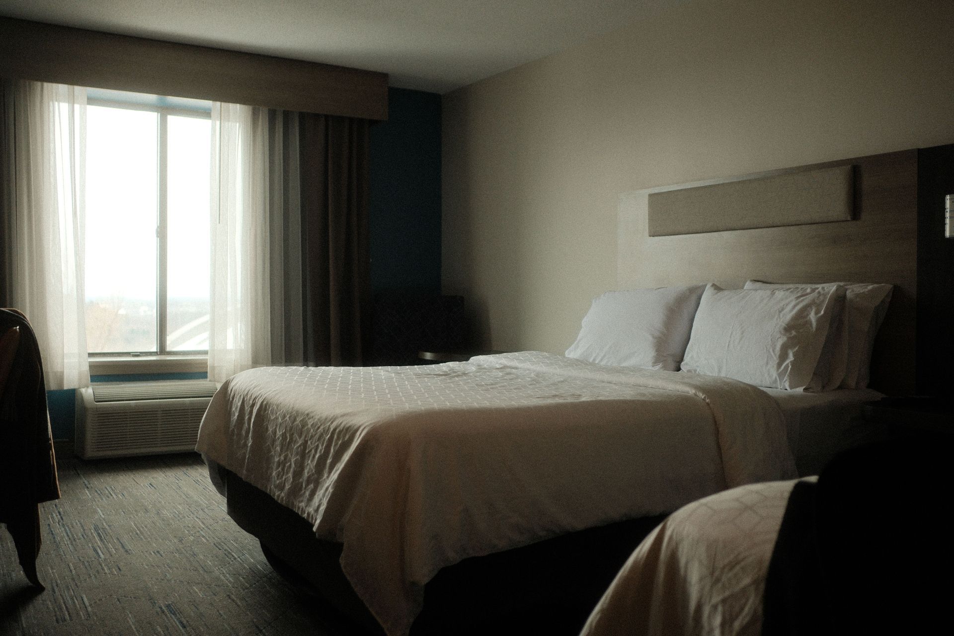 Dim hotel room with two white beds, window curtains, and an air conditioner by the window.
