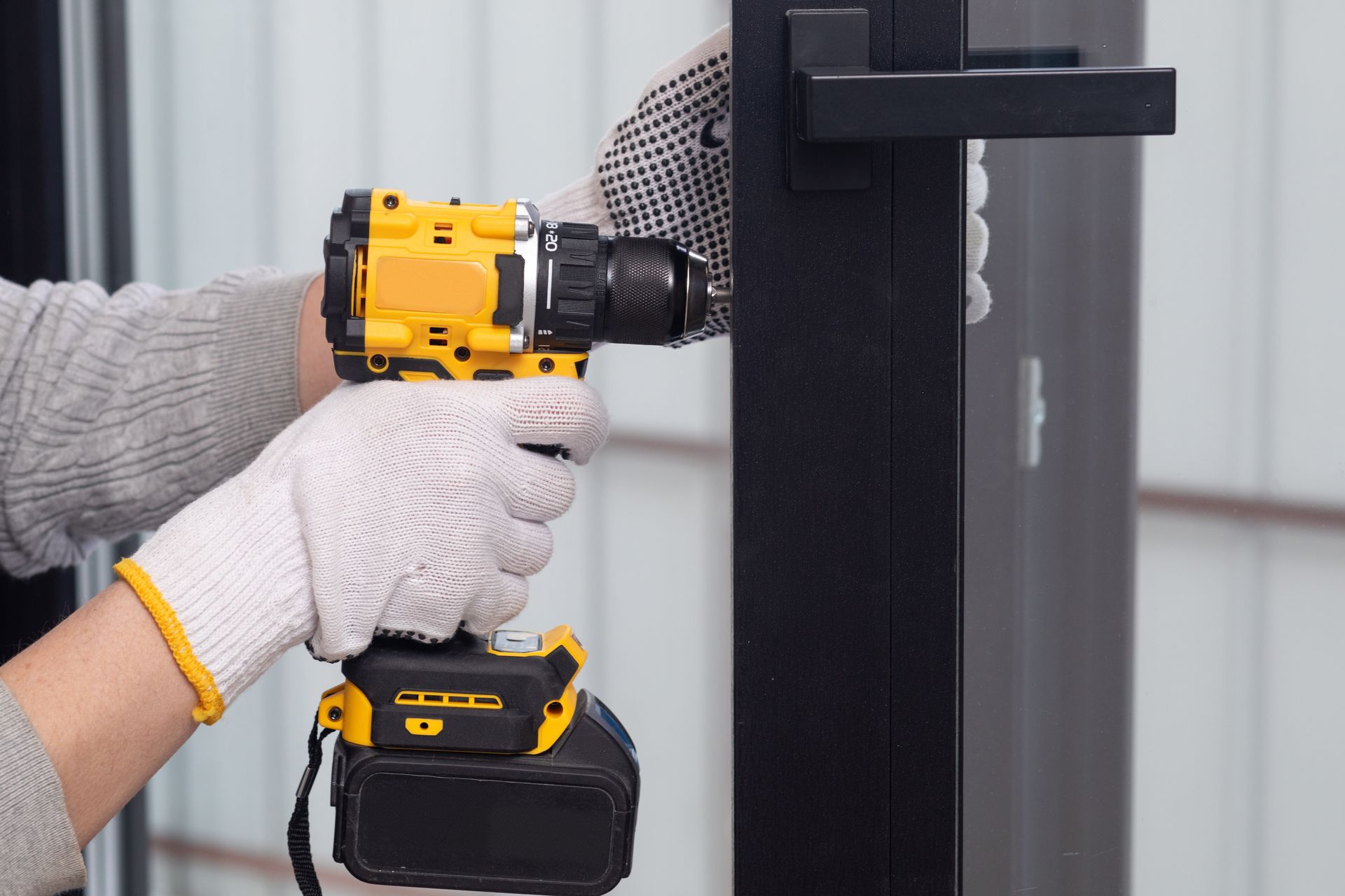 Person using a yellow drill to install a black door handle, wearing gloves.
