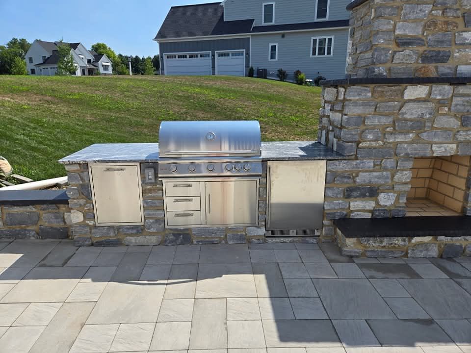 Jesse James Hardscaping - Outdoor kitchen with stone façade, stainless steel grill, cabinets, and fireplace, on a patio.