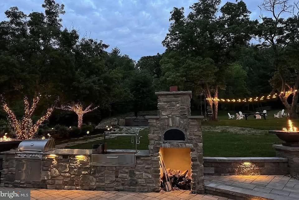 Jesse James Hardscaping - Backyard patio with outdoor kitchen and wood-fired oven at dusk, string lights, trees with lights, and a lawn.