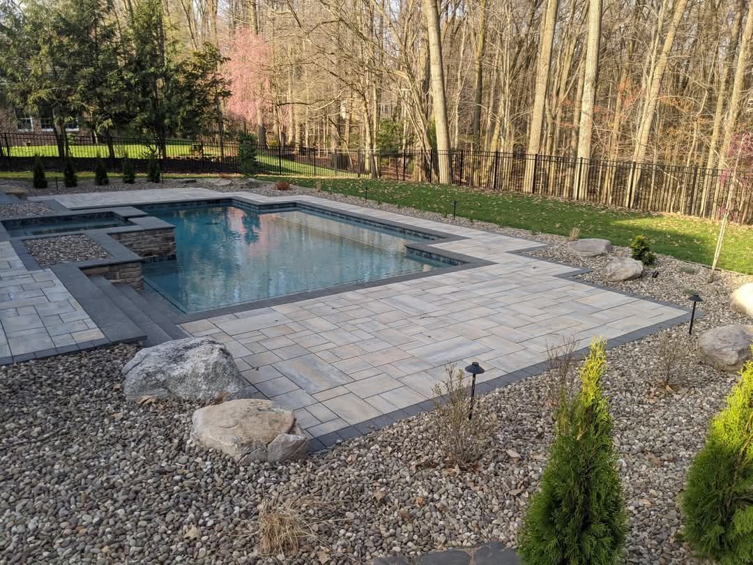 Jesse James Hardscaping - A modern pool with stone patio, surrounded by trees, grass, and landscaping.