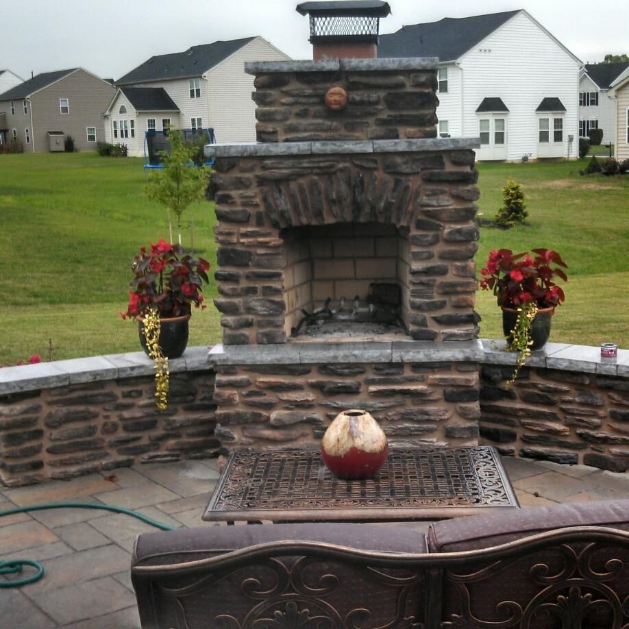 Jesse James Hardscaping - Outdoor stone fireplace with seating, potted red flowers. Backyard setting, houses in the background.