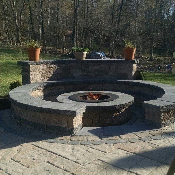 Jesse James Hardscaping - Circular fire pit made of stone with flames, surrounded by seating and potted plants in a backyard.