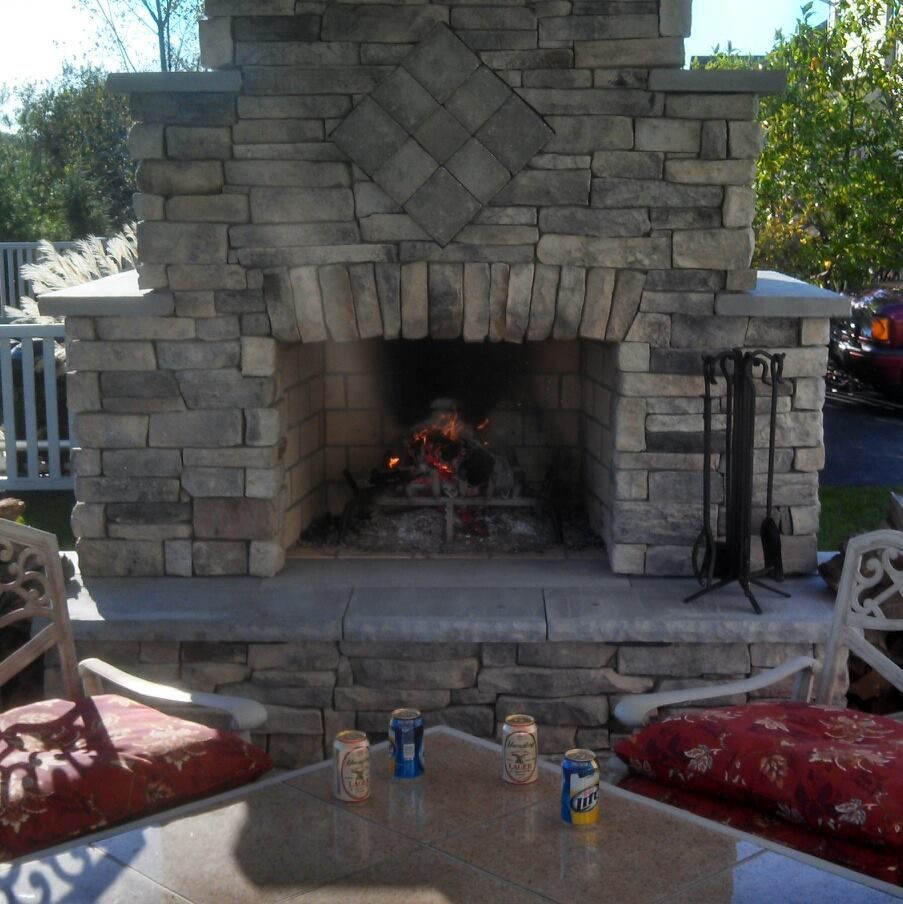 Jesse James Hardscaping - Stone outdoor fireplace with a fire burning, surrounded by seating and a table with drinks.