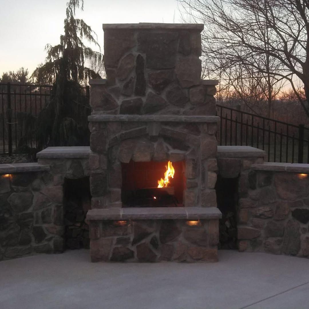 Jesse James Hardscaping - Stone outdoor fireplace with lit fire, flanked by low walls, evening setting.