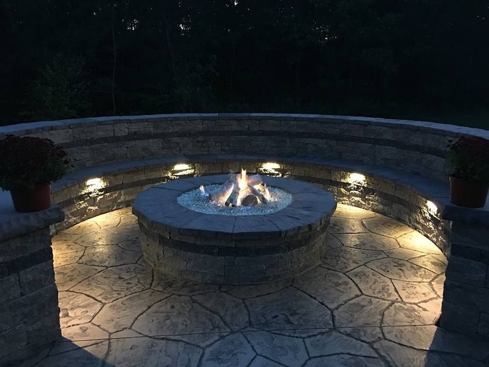 Jesse James Hardscaping - Fire pit in circular stone patio, surrounded by tiered seating and lights at dusk.