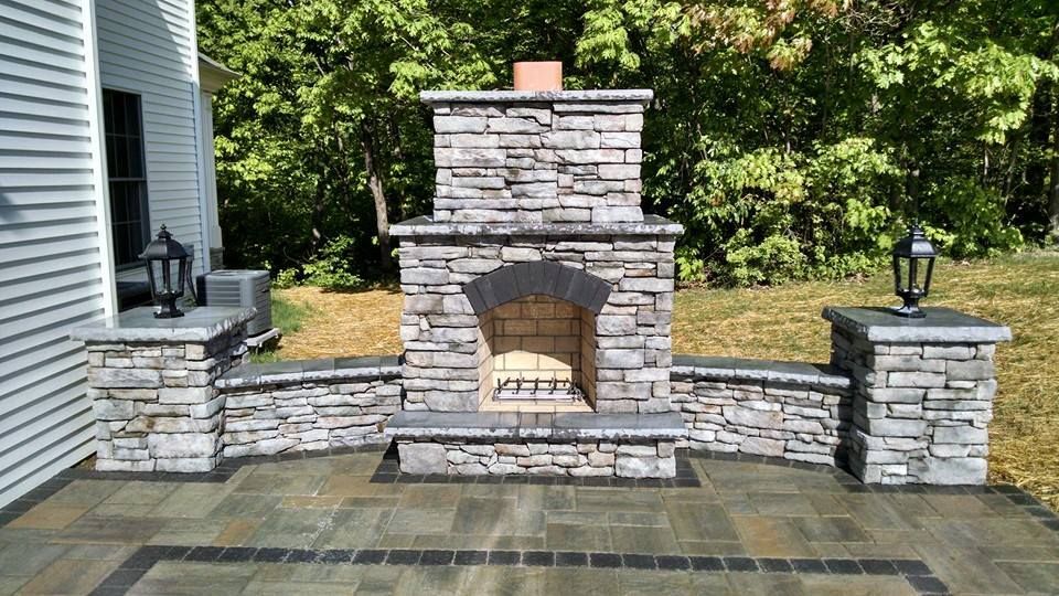 Jesse James Hardscaping - Outdoor stone fireplace with flanking pillars, on a paved patio, set against a backdrop of trees.