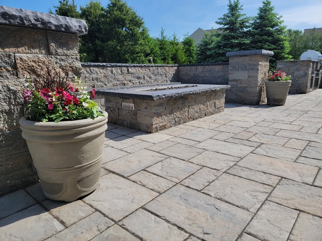 Jesse James Hardscaping - Stone patio with planters, a fire pit, and a low wall on a sunny day.