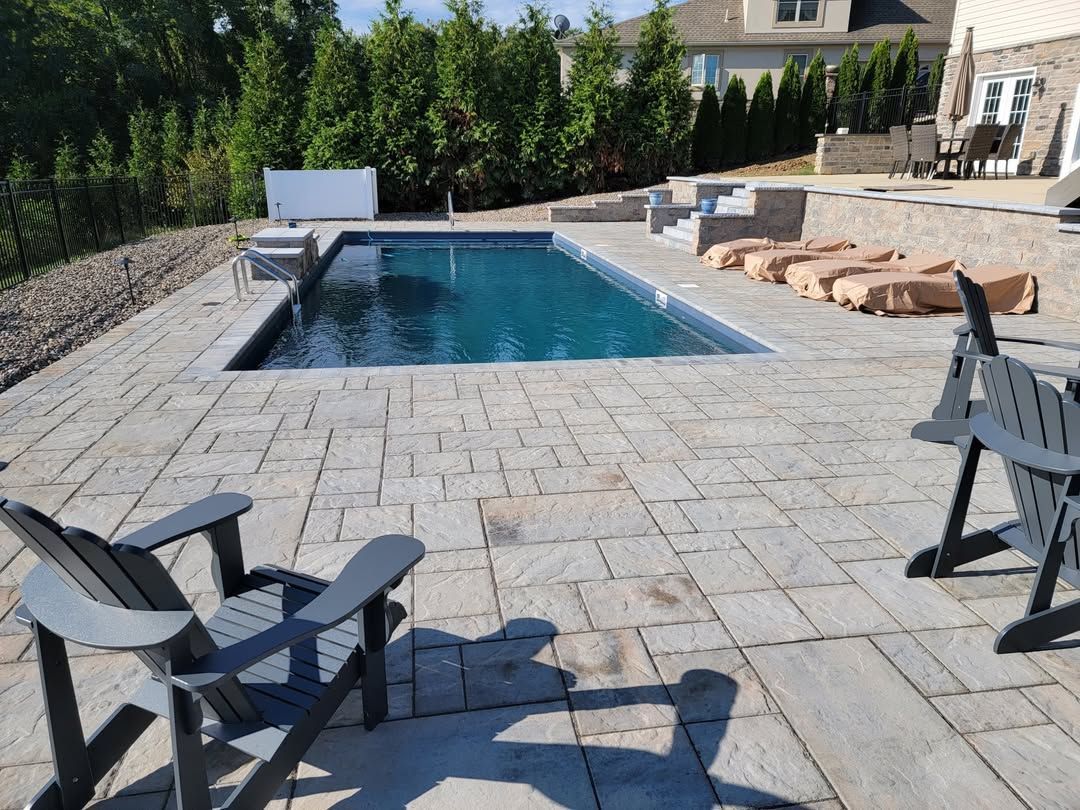 Jesse James Hardscaping - A backyard pool with a paved patio, two gray Adirondack chairs, and lush greenery.