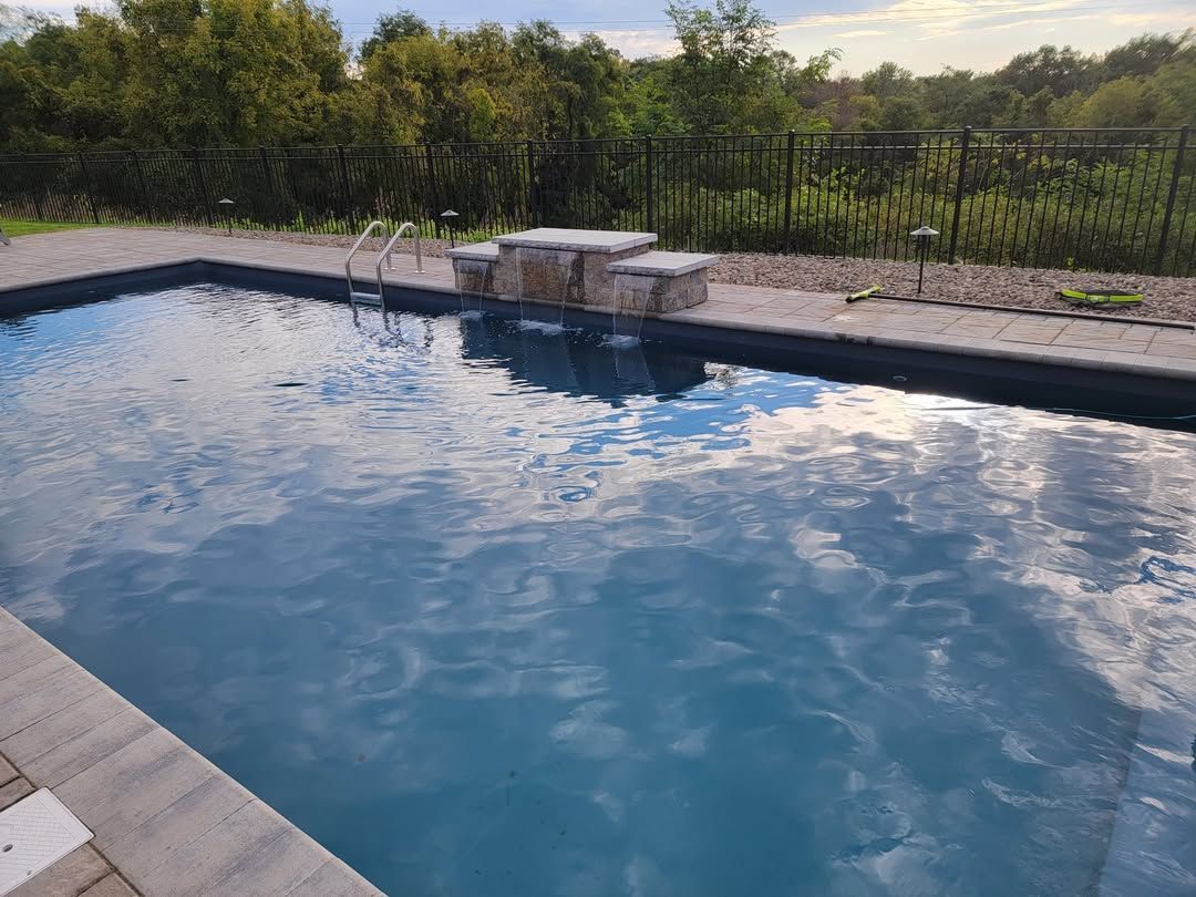 Jesse James Hardscaping - A rectangular pool with stone surround and waterfall feature, reflecting a cloudy sky.
