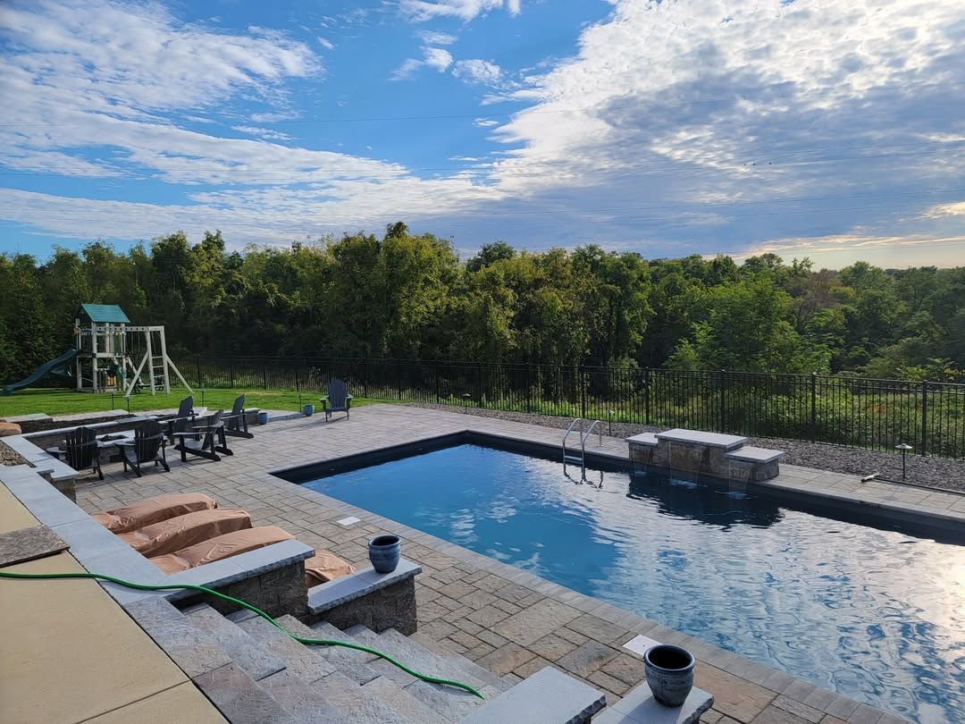 Jesse James Hardscaping - A rectangular pool with a view of trees, a playset, and a blue sky with clouds.